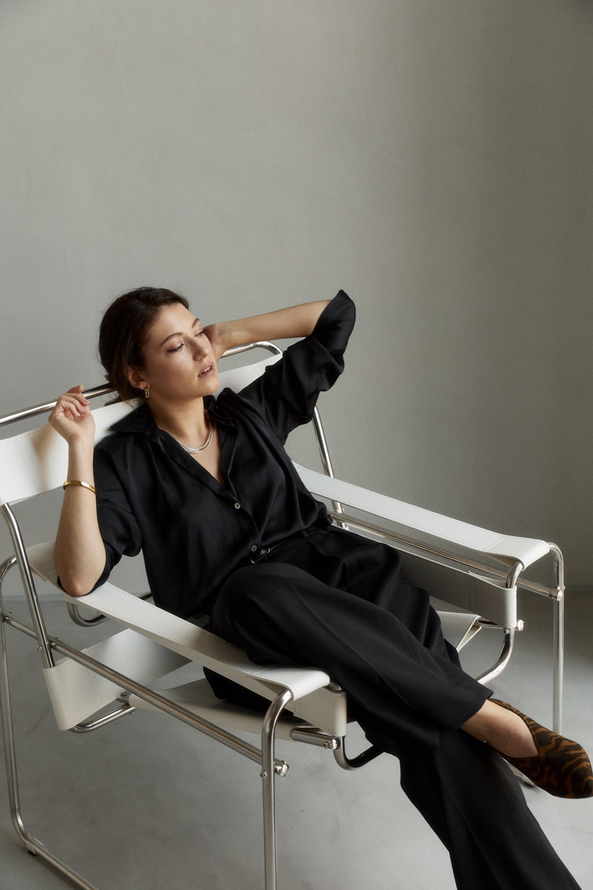 Ksenia in black, reclining in a chrome and white chair with one hand at her hair