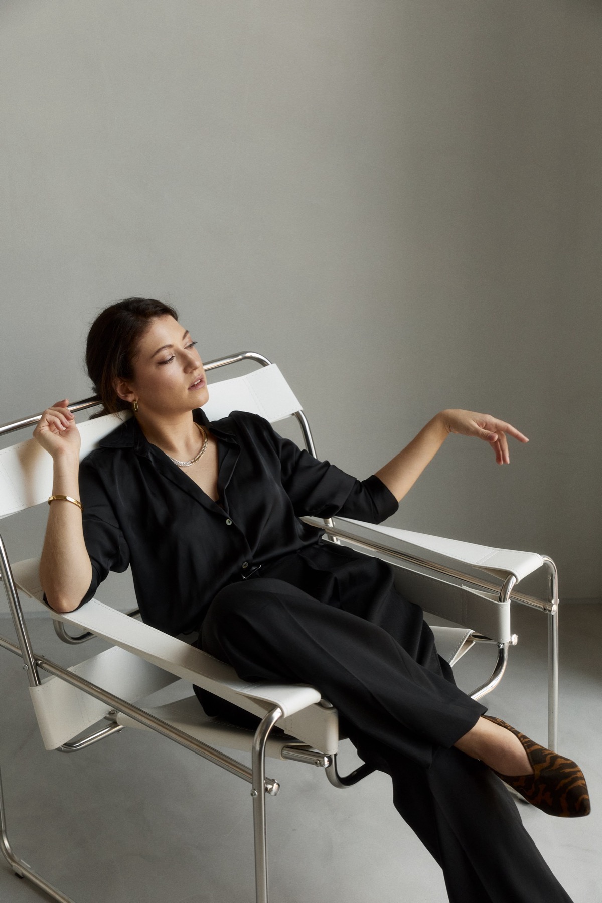 Ksenia in black, seated in a chrome and white chair, looking away from the camera